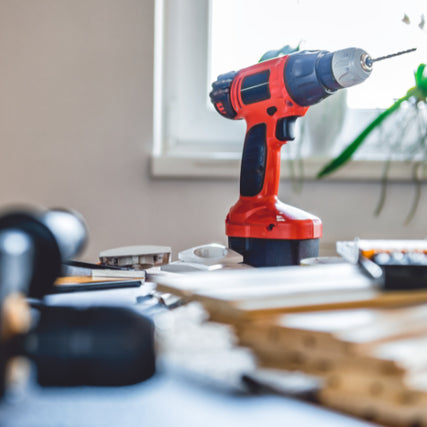 Power ToolsA red power drill stands upright on a cluttered workbench with scattered tools and papers in a sunlit room, creating a busy, industrious atmosphere.