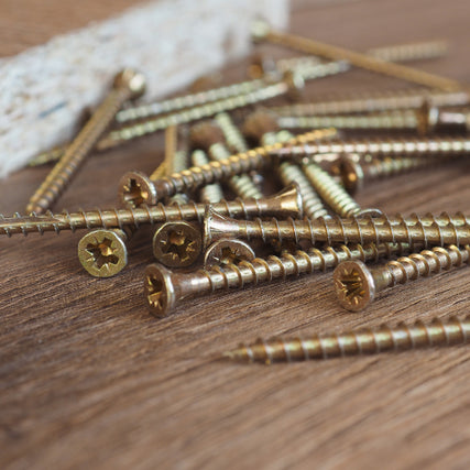 HardwareA scattered pile of brass screws on a wooden surface, with a blurred wooden board in the background. The image conveys a workshop or DIY setting.