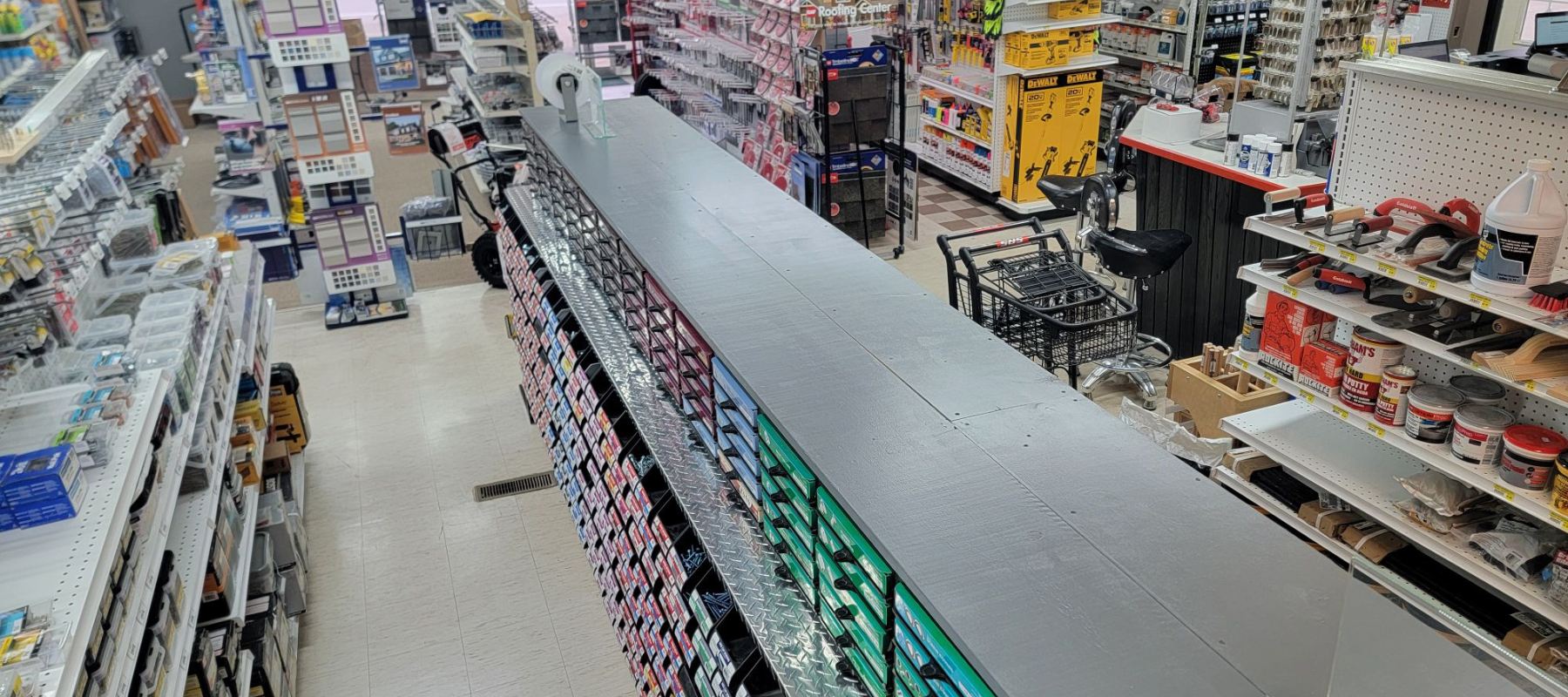 Overhead view of a hardware store aisle showing shelves stocked with tools, paint cans, and supplies. The setting is bright and organized.